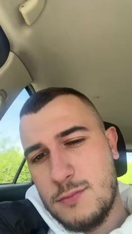 The video captures a car journey in Voysil, Bulgaria, during the daytime. The camera, likely held by the man, is positioned in the front passenger seat.

Initially, a close-up shows the man's face, looking downwards, with a short haircut and light stubble. The camera then pans to the right, revealing a woman in the driver's seat. She wears sunglasses, and her dark hair is pulled back. The car's interior, including the headrest, dashboard, and the side window, is visible.

The camera continues to pan towards the open car window, showcasing a clear blue sky and vibrant green trees outside, indicating bright and sunny weather. A seatbelt mechanism on the car's B-pillar is also visible. Finally, the camera pans back, showing a partial view of the man's face, with the blue sky still visible through the window above. The overall scene depicts two individuals driving in a car on a pleasant day. No text is visible in the video.