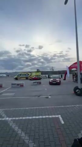 The video shows a red Subaru Impreza WRX hatchback driving around an obstacle course. The car is driving on a paved surface with white painted parking lines. There are concrete block barriers and white flags marking the course. An ambulance and other vehicles are parked in the background. The sky is cloudy and overcast. A person in a dark jacket can be seen watching the car.