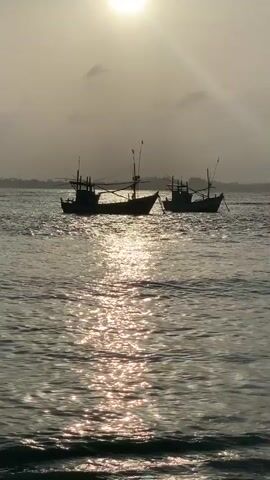 Two fishing boats are silhouetted against the sun on the water. The sun is a bright orb in the sky, partially obscured by clouds. The water is choppy, with sunlight reflecting off the waves. The boats are small and traditional, with tall masts. The overall mood is serene and peaceful.