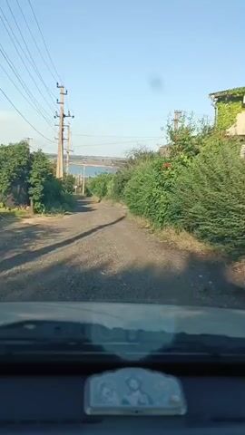 A first-person view from inside a vehicle shows a gravel road winding uphill. On either side of the road, there is dense green foliage and several utility poles with wires stretch across the frame. The road continues upwards, curving to the right, and in the distance, a body of water can be seen under a clear blue sky. The dashboard of the vehicle is visible at the bottom of the frame, with a small religious icon placed on it. The video appears to be taken during daylight, with the sun casting shadows on the road.