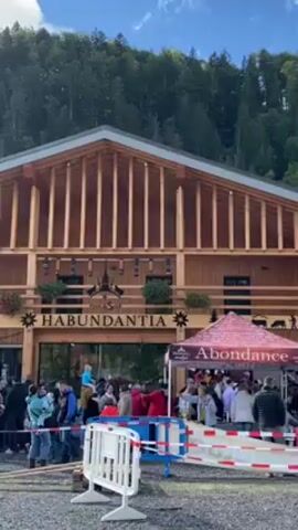 The video shows a crowd of people gathered outside a large wooden building with the name "HABUNDANTIA" above the entrance. The building is set against a backdrop of a lush green hillside and a partly cloudy sky. There are barriers and tents set up near the entrance, likely for crowd control or event purposes. People of various ages and demographics are visible, some walking, some standing, and some carrying children on their shoulders. The atmosphere appears lively and possibly festive. The presence of the crowd and the decorations suggest a public event or gathering.