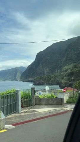 The video presents a landscape view from a vehicle, likely a car, showing a coastal area with mountains in the background. The sky is partly cloudy. The foreground is dominated by a road with a pedestrian crossing, and on the side of the road there is a small building or structure with a "no entry" sign posted on it. To the left, there's a gated entrance, possibly to a private property. The mountains are lush with green vegetation, and the sea is visible in the distance. There are no people visible in the frame. The camera is static, providing a still view of the landscape.