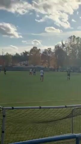 The video shows a soccer match in progress on a grassy field on a sunny day with scattered clouds in the sky. Players in red and white uniforms are visible, along with players in black and white uniforms, running across the field. A referee in a green uniform is also present. The camera is positioned behind a soccer goal, and the net is visible in the foreground.  Trees with autumn foliage line the perimeter of the field. A building is visible in the background.