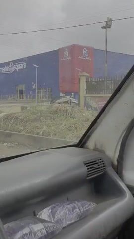 The video provides a first-person perspective from inside a vehicle, looking out through the windshield and side windows. The exterior scene shows a large, modern building with a distinctive blue and red facade. The building has prominent signage that reads "Market Square" and "Fashion World." Several streetlights are visible in front of the building. There are a few vehicles parked in front of the store, including a silver SUV and a dark car. In the distance, a few people can be seen on the premises of the store, some appearing to be workers engaged in activity. The surrounding environment includes a paved area, some overgrown vegetation, and a drainage grate. The sky is overcast, suggesting a cloudy day. The interior of the vehicle is partially visible, showing parts of the dashboard and a door panel. The video captures a brief moment of passing by the building.