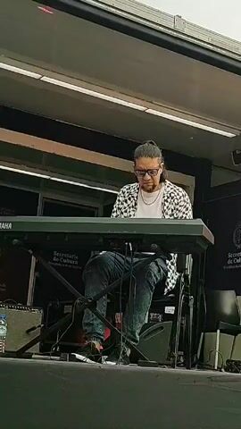 A man wearing sunglasses, a white t-shirt, and a patterned jacket is seated on a stage, playing a Yamaha keyboard. He has long hair tied back and wears jeans and dark sneakers.  The keyboard stand is labeled "Secretaria de Cultura". An amplifier and a water bottle are visible on the floor to his left. A black chair and another sign for "Secretaria de Cultura" are to his right. The stage is under an awning.