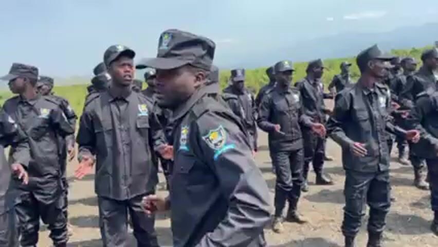 Here is a description of the video:

The video shows a large group of men in black uniforms and caps clapping and singing together outdoors. They stand in rows against a backdrop of green fields and distant mountains under a bright sky. The uniforms have patches on the shoulders. The men seem to be performing or practicing some kind of synchronized activity, likely a form of song and dance. The overall tone of the scene is spirited and unified.