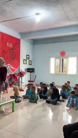 The video shows a group of young children and a few adults in a room, likely a classroom or community center. The children, dressed in blue shirts and some in headscarves, are sitting on the floor in neat rows, facing towards the left side of the frame. They appear to be engaged in some sort of activity or lesson.

The room has light blue walls and a white ceiling with visible fluorescent lighting. There is a large window on the right wall, letting in natural light, suggesting it is daytime. The floor is tiled and reflective. On the left wall, there is a red mural with musical notes and the word "Music" written on it, along with some abstract geometric shapes. A speaker and a red chair are also visible near this wall. A clock with a red face is mounted on the wall above the window.

The adults, also in modest attire, are seated or standing, observing the children. One adult in a purple headscarf and dress is seated in the foreground on the left, looking down at her phone. Another adult in a similar headscarf is seated on the right. The overall atmosphere seems calm and attentive. The children's faces are mostly turned away from the camera, but their posture suggests they are focused on something or someone to their left, out of frame. There are no strong indications of specific emotions, but the scene implies a structured learning or entertainment environment for children.

The location context provided is Tanjung Gading, Indonesia.
