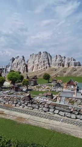 A miniature model village is shown set against a backdrop of large, rocky mountains. In the foreground, there are model train tracks with a grassy area on either side. Several small, model buildings are scattered throughout the village, including a church with a tall steeple. A winding road with two model cars is visible in the mid-ground, leading towards the mountains. The sky is partly cloudy. The scene appears to be outdoors, under natural light. There are no people visible in the video.