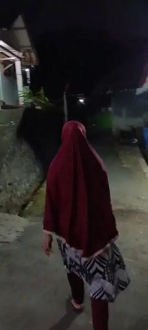 A person wearing a dark red headscarf and a patterned skirt walks away from the camera down a paved alley at night. The person is walking on the right side of the alley. On the left side of the alley is a raised concrete embankment with some vegetation. Buildings with lights are visible on either side of the alley in the background. Towards the end of the alley, a motorcycle with its brake lights on is visible. The person is walking towards the motorcycle. The lighting is dim, with artificial lights illuminating parts of the scene. The person's feet are obscured by their skirt.