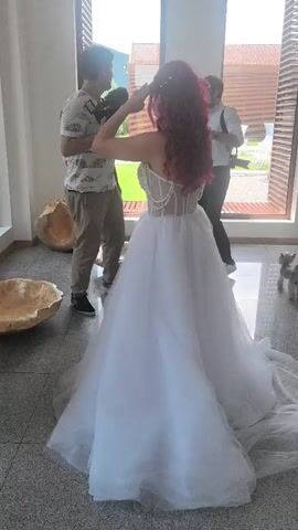 A woman in a white wedding gown, with reddish-pink hair, adjusts her hair while facing away from the camera. To her left, a man in a white and black patterned shirt and khaki pants holds a camera and observes her. To her right, another man in a white shirt and suspenders holds a camera and looks towards her. In the background, large windows reveal an outdoor scene with green hills and modern buildings. Two small dog sculptures are visible near the windows. On the left side of the frame, large, organic-shaped objects in a light brown color are on the floor. The floor is tiled in a light gray color.