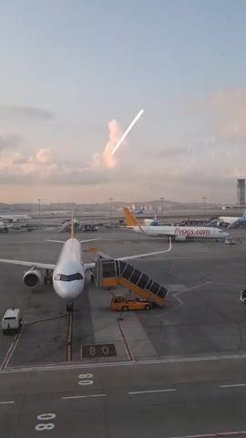 The video shows an airport tarmac at sunset. Several airplanes are parked, with one in the foreground connected to a mobile staircase. A small vehicle is positioned near the front of this aircraft. In the background, more planes are visible, along with a distant cityscape. The sky is filled with clouds tinged with the colors of sunset. A streak of light, possibly from a plane in flight or a lens flare, is visible in the upper portion of the frame. The overall impression is one of a busy airport at the end of the day.