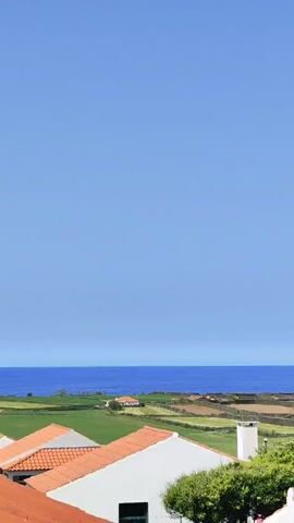 The video opens with a panoramic view of a vibrant coastal landscape under a clear blue sky. In the foreground, red-tiled rooftops of several white buildings are visible, including a prominent white chimney. Beyond the rooftops, expansive green fields, divided by numerous low stone walls, stretch towards the horizon. The deep blue ocean forms a striking line against the sky.

As the camera slowly pans from left to right, more of the picturesque scenery is revealed. The initial view shows a vast expanse of ocean and farmland. Towards the middle of the pan, a small, densely vegetated area with trees breaks up the fields. Finally, a charming coastal town with white houses and red roofs nestled along the shoreline comes into view on the right side of the frame, hinting at a more populated area. The overall impression is one of a serene and sunny day in a beautiful rural coastal setting. No people or visible text are present in the video.