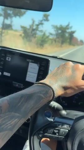 A first-person perspective video shows a hand with tattoos gripping a steering wheel of a Toyota car. The dashboard displays speed and fuel information. The car is driving on a road lined with pink flowering bushes under a clear blue sky. In the distance, the ocean is visible.