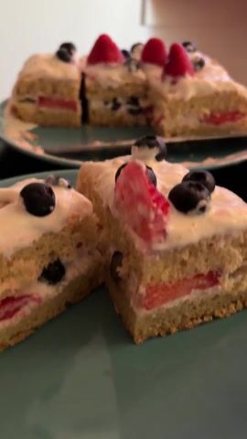 The video opens with a close up of a plate of cake, with two slices on another plate in the foreground. The cake is a light color, frosted with white icing and topped with blueberries and sliced strawberries. The cake is layered, with strawberries and blueberries in between the layers. The camera then pans up to show a TV screen playing a scene from a TV show, with a woman with long, dark hair wearing a light purple shirt. The TV is on a light-colored stand, with red wall behind it. There is a flower on top of the stand to the left. The screen has Chinese subtitles.