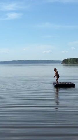 A young person stands on a black inflatable raft in the middle of a lake under a blue sky with sparse clouds. The person, wearing dark swimwear, is fair-skinned with short blond hair. The water is calm, with subtle ripples creating a horizontal lined pattern.  In the background, there is a landmass with a green forest. The person bends their knees, leans forward, and jumps off the raft into the water. The raft tips slightly as they jump. A small splash marks the spot where they enter the water.