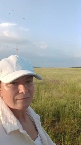 A woman wearing a white baseball cap and a white shirt is seen from the chest up, facing to the right. She is in a grassy field under a light blue sky with some clouds. There are several small dark objects, possibly insects, in the sky in the upper left portion of the frame. A utility pole is visible in the background to the left. The woman appears to be looking towards the field. The lighting suggests it is daytime, possibly late afternoon or early morning due to the soft light.