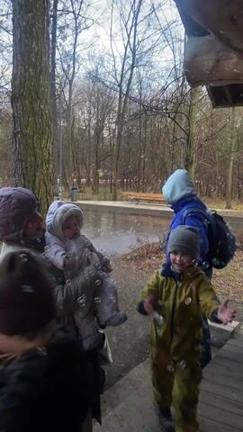 The video depicts a group of people, including adults and children, in what appears to be a park or wooded area on an overcast day. There are bare trees in the background and a body of water visible. A woman is holding a baby in her arms. The children are playing, and a person is blowing bubbles, filling the air with them. People are wearing coats and hats, suggesting a cool temperature. Some individuals are walking, while others are standing and interacting. The general atmosphere is playful and festive.