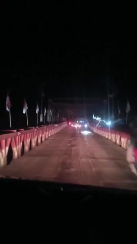 The video is a first-person perspective shot from inside a vehicle driving across a bridge at night. The bridge has red and white striped barriers along the sides, with flags mounted on poles. There are streetlights and vehicle headlights visible on the bridge, creating streaks of light due to the motion. The overall scene is dark with limited visibility beyond the bridge itself.