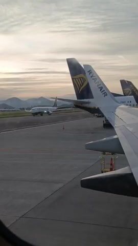 The video captures an airport scene, likely at Orio al Serio, Italy, during early morning or late afternoon, characterized by soft, warm light and a partly cloudy sky. From a window perspective, the wing and tail fin of a Ryanair airplane are prominently featured in the foreground. The tail displays the "RYANAIR" logo and the partial registration "I-DWG".

In the mid-ground, another Ryanair airplane is seen taxiing across the tarmac, with its navigation lights illuminated. "RYANAIR" is also visible on its fuselage. Mountains form a scenic backdrop in the distance. Additional Ryanair aircraft tails are visible further in the background. The tarmac is clear, with a few small orange safety cones visible. The scene conveys an active airport environment at a calm, visually appealing time of day.