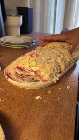 A close-up shot focuses on a person's hands slicing a meringue roll dessert on a wooden table. The dessert is elongated and covered in a cracked, light-colored meringue shell, with visible layers of cream and red berry filling. The person is using a large metal knife to cut the dessert, and small pieces of meringue are scattered on the table. In the background, there are stacks of plates and a blurred out interior. The lighting is bright and appears to be natural.