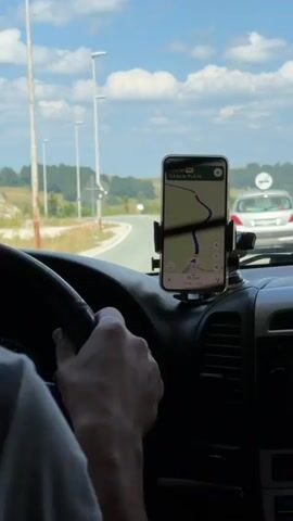 The video shows a first-person perspective from inside a vehicle driving on a road. A hand is visible on the steering wheel. A smartphone is mounted on the dashboard, displaying a navigation app with a map and route information. The road ahead curves to the left, and a silver car is visible in front. The surrounding environment appears to be rural, with rolling hills, trees, and a blue sky with clouds. There are streetlights visible along the side of the road in the initial frames.