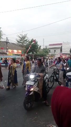 The video shows a street scene with people participating in what appears to be a parade or procession. In the foreground, a man is riding a motorcycle. To his right, a woman is riding a bicycle. Behind them, a group of people are walking, some in traditional clothing. There are also shops and buildings lining the street, and Indonesian flags are visible. The sky is overcast.