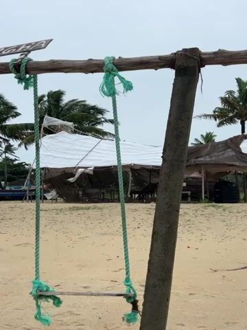 The video shows a close-up view of a swing made of rope and a wooden seat, hanging from a wooden beam supported by two posts. The swing is on a sandy beach. In the background, there are several makeshift structures with white and brown tarps, likely serving as shelters or stalls. Palm trees are visible behind these structures, and the sky is overcast. The overall impression is of a beachside setting, possibly a tourist spot, with a focus on the simple swing.