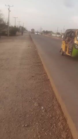 The video shows a road with a dirt shoulder on the left. Several vehicles, including a silver sedan, a yellow auto rickshaw, a motorcycle, and a large truck, are moving on the road. There's a person walking in the distance on the left side of the road. Power lines are visible on the left side, and buildings are seen on the right side of the road. The sky is overcast.