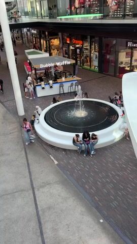 This is a high-angle, slightly shaky video taken inside a modern indoor shopping mall. The camera pans slowly to the right. In the foreground, a circular white bench surrounds a dark water fountain with a spouting water feature. Several people are seated on the bench, mostly young women, some looking at their phones. To the left of the fountain, a woman walks by carrying a large tote bag. Further back, a kiosk labeled "Bubble Waffle" is visible, with staff serving customers and more people standing around. In the background, shops with large glass windows line the mall corridor, displaying merchandise and store names like "Snipes," "Orange," and "Histoire." Sunlight streams in from above, creating bright patches and reflections on the floor. The overall atmosphere appears to be a bustling, casual day at a shopping center.