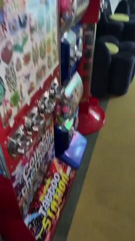The video shows a close-up of a vending machine labeled "Tattoo Station". The machine is red, blue, and yellow, dispensing temporary tattoos. It appears to be located in a public area, possibly a waiting room, given the presence of dark blue and green upholstered chairs in the background. The camera is moving slightly. The vending machine has several dispensing slots, and a sticker-covered panel on its side.