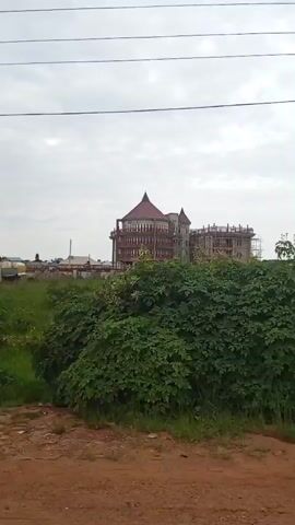 The video shows a construction site with a large building under construction. The building is multi-story with a distinctive conical roof. In the foreground, there are stacks of bricks and other building materials. Vegetation, including bushes and grass, is visible throughout the scene. There are also power lines and a utility pole. The ground is a mix of dirt and gravel. The sky is overcast. The camera is moving, showing different angles of the construction site. No people are clearly visible in the video.