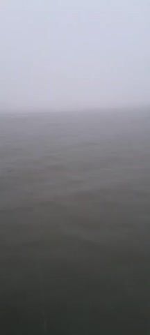 A hazy, gray sky meets a body of water that appears to be choppy. There is evidence of light rain falling on the water's surface, visible as streaks. The scene is devoid of any discernible objects or people, suggesting a vast, open expanse of water under overcast conditions. The camera angle is from a high vantage point looking down onto the water.