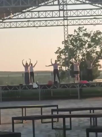 The video shows an outdoor stage at sunset. On the stage, five people are visible, appearing to be practicing a dance routine. They are positioned at various points on the stage, some with arms outstretched. In the foreground, several empty benches or tables are visible, suggesting an outdoor seating area. The background is a blurry landscape with trees and a structure resembling a large canopy or roof over the stage. The lighting suggests it is either early morning or late evening.