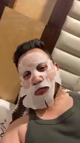 A person wearing a white sheet mask is lying down. The person has dark hair and a beard and is wearing a green tank top and a gold chain. Someone with a tattoo on their arm is adjusting the mask. The person is smiling slightly. The background includes a tan headboard and possibly a wall.