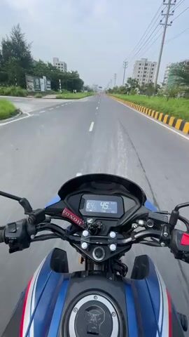 The video is filmed from the perspective of someone riding a blue and white Honda motorcycle. The camera is mounted on the handlebars, showing the digital speedometer which indicates a speed of 45 km/h at the beginning of the clip, gradually increasing to 48 km/h by the end. The rider's hands are visible on the handlebars, operating the controls. The road ahead is paved with asphalt, marked with white lines and bordered by a yellow and black striped barrier on the right side. On the left side of the road, there is a grassy area with trees and some buildings in the background. On the right side, there are also buildings and utility poles with overhead wires. The sky is overcast. There are orange traffic cones visible on the road in the distance, suggesting construction or a diversion. The motorcycle has a prominent Honda logo on its fuel tank. No other people are clearly visible in the video. The environment appears to be an urban or suburban area, possibly on the outskirts of a city.