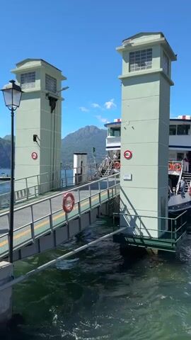 The video shows a ferry docked at a pier with two distinctive pastel green towers. A gangway extends from the pier to the ferry, and several people are visible on the gangway and the deck of the ferry. The water is clear and green, with small waves. In the background, a mountain looms under a clear blue sky. The ferry appears to be a passenger vessel, with windows along its upper deck. Life preservers are visible on the gangway and the ferry. There are signs with red circles and numbers on the towers and the ferry, likely indicating speed limits or other regulations. The lighting suggests it is a sunny day.