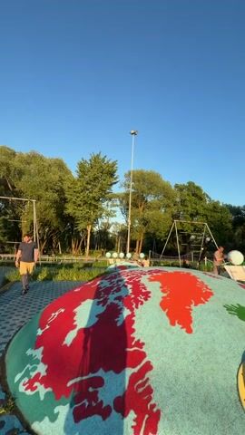 The video shows a children's playground with various play structures. The scene is outside on a sunny day with some clouds in the sky. 

Initially, the camera focuses on a dome-shaped structure painted with a world map. The camera pans across the playground, showing swings, other children's play equipment, and people, including children and adults. Some people are sitting on the swings, while others are walking around. The ground surface varies, including blue patterned bricks and grassy areas. In the background, there are trees and a building that appears to be a commercial or public structure.