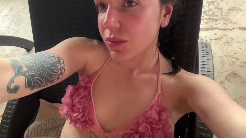 A woman in a pink and blue bikini is reclining on a black patio chair. She has dark hair, fair skin, and a tattoo on her left arm. She is taking a selfie. The background is blurry, but it appears to be an outdoor area with tile flooring.