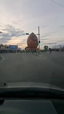 The video shows a time-lapse of a traffic circle at dusk. A large, brown, stylized nut-shaped monument is visible in the center of the circle. Traffic lights are also present and illuminated. The sky is cloudy with hints of orange and purple from the setting sun. The surrounding area appears to be a town or city with buildings and some trees visible. The perspective is from inside a vehicle, looking forward.