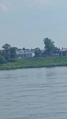 The video shows a wide, calm river with gentle ripples on its surface. The camera is positioned on the water, looking towards the opposite bank. The bank is lined with lush green vegetation, including trees and bushes. In the background, there are several buildings of varying sizes and architectural styles, some appearing to be houses and others possibly part of a village or town. One prominent building on the bank has a distinctive white structure with a blue dome and spire, resembling a church or a monastery. The sky above is partly cloudy and appears to be a bright, clear day. The video is a continuous shot, panning slightly to the left as it progresses, revealing more of the landscape and buildings along the riverbank. There are no people or animals visible in the footage.