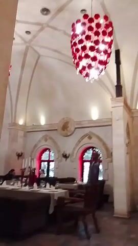 The video shows the interior of a restaurant. The room has a high, arched ceiling, with a large red and white chandelier hanging from it. There are arched windows with red trim around them. Tables with white tablecloths and grey chairs are arranged throughout the room. People are seated at some of the tables. A balcony is visible at the back of the room. The overall style of the restaurant is somewhat gothic or medieval.