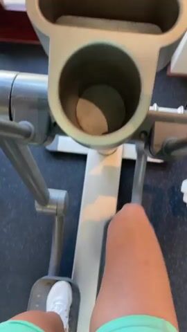 The video shows a first-person perspective of someone exercising on an elliptical machine. The machine is light-colored with gray accents. The person is wearing white sneakers and teal shorts. The console of the elliptical is visible, displaying some text and indicators. In the background, through a window, there is a view of a swimming pool, palm trees, and buildings. The scene appears to be outdoors in a sunny location, possibly a resort or a gym with a view. The overall atmosphere is one of fitness and vacation.
