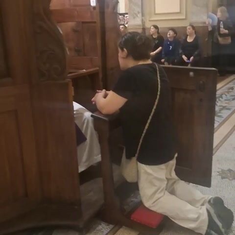 The video shows a woman kneeling in a confessional booth in a church, speaking to a priest. The woman is wearing a black t-shirt and light-colored pants, with a purse slung over her shoulder. She is kneeling on a red cushion and resting her arms on the ledge of the booth. The priest is visible through a screen. He is wearing a white clerical collar and a purple stole. He is leaning forward and listening to the woman. Other people are sitting in the pews in the background. The floor appears to be mosaic. The confessional booth is made of dark wood.