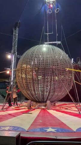 A medium shot depicts a circus setup under a blue tent. Two men are seen near a large, spherical metal cage supported by a wheeled base and vertical stands. The cage has a grid-like pattern and a light inside. Above the cage, gymnastic rings hang from a suspension rig. The circus ring features a red and white striped pattern with blue accents and stars. Additional lighting structures and rigging are visible around the periphery.