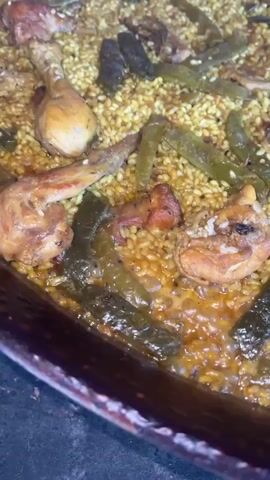 The video shows a close-up of a large pan of paella cooking on a stove. The paella includes rice, pieces of chicken, and green beans. The rice is yellow and slightly soupy. The chicken pieces are brown and appear cooked. The green beans are long and dark green. The pan is circular and has a dark rim. The stove appears to be indoors with tiled walls. The camera moves around the pan slightly to show different parts of the paella.