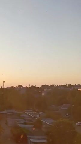The video shows a time-lapse of a sunrise over a suburban or semi-urban area. The sun is visible as a bright orb, gradually rising and casting a warm, golden light across the scene. Silhouetted trees and buildings are visible in the foreground and midground. The sky transitions from a pale yellow near the horizon to a soft blue overhead. There are some blurry circular light artifacts throughout the video, likely lens flares or dust on the camera lens. The image is vertically oriented.