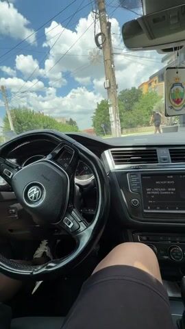 This video shows the interior of a car, likely a Volkswagen, from the perspective of the driver. The steering wheel, dashboard, and infotainment system are visible. The car appears to be stationary, with the engine on, as indicated by the dashboard lights. Outside the car, a man is walking on a sidewalk next to a street. The weather appears to be sunny with some clouds.