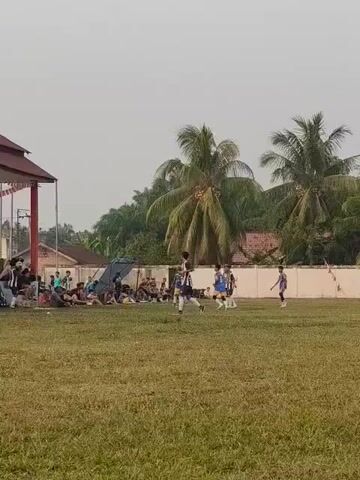 A wide shot captures a soccer game played on a grassy field under a hazy sky. Several players in matching uniforms, some blue and white, others black and white, are scattered across the field, actively engaged in the match. Spectators sit on the grass and under a covered structure on the left side of the frame, watching the game. Palm trees and other foliage line the background, behind a low wall.