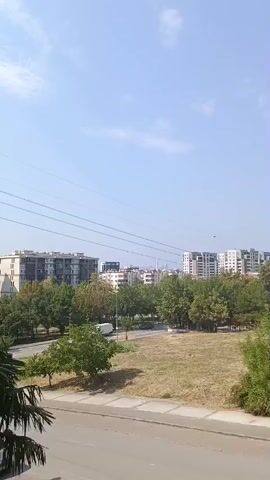 A slightly shaky video pans across a dry, grassy field with scattered trees and bushes. In the background, several multi-story apartment buildings are visible, some with balconies. A road with a white van driving on it is present to the left of the field, and overhead power lines are strung across the sky. The sky is clear and blue with a few wispy clouds.