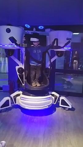 A small boy stands inside a large white and black VR simulator in a room with blue lighting. He is wearing a blue shirt with "ITALIA" written on it, brown pants, and a VR headset. He extends his arms out to the sides, holding onto black handles inside the simulator. The simulator rotates during the video, allowing a view of its various components. The room has wood-look flooring, and there is a woman visible behind a glass door in the background. A fire extinguisher is visible on the wall near the door. The room appears to be some kind of entertainment or arcade space. The simulator has blue lights around its base.